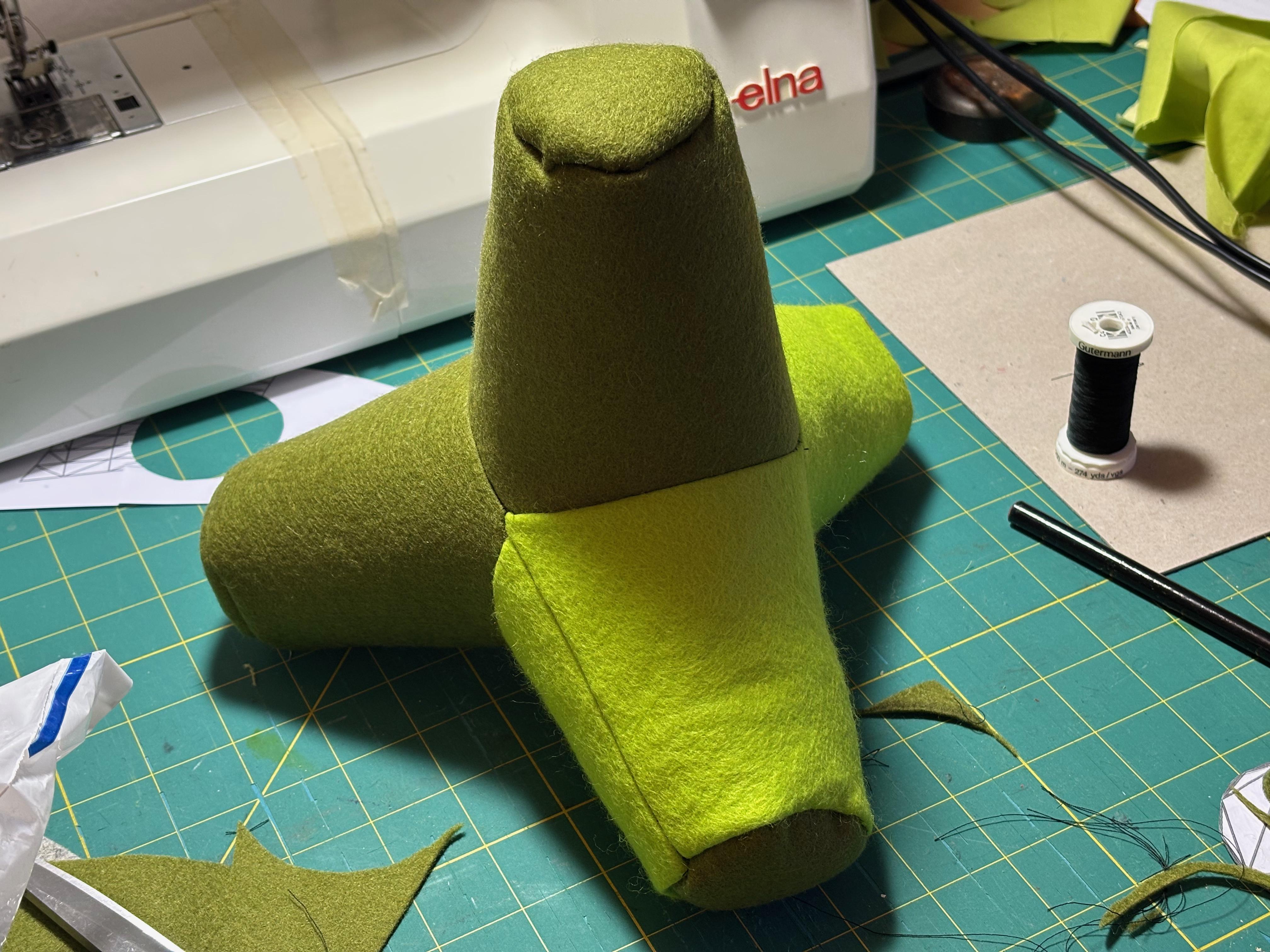 A tetrapod (4-legged geometric shape) made out of two shades of green felt resting on a cutting mat, surrounded by sewing tools, thread, offcuts and a sewing machine