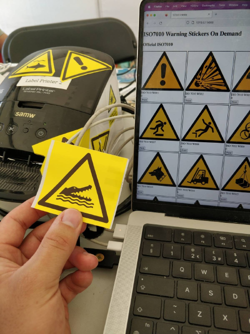 A photo of a hand holding a crocodile warning sticker. In the background is a laptop showing a web interface with a grid of warning signs and a label printer