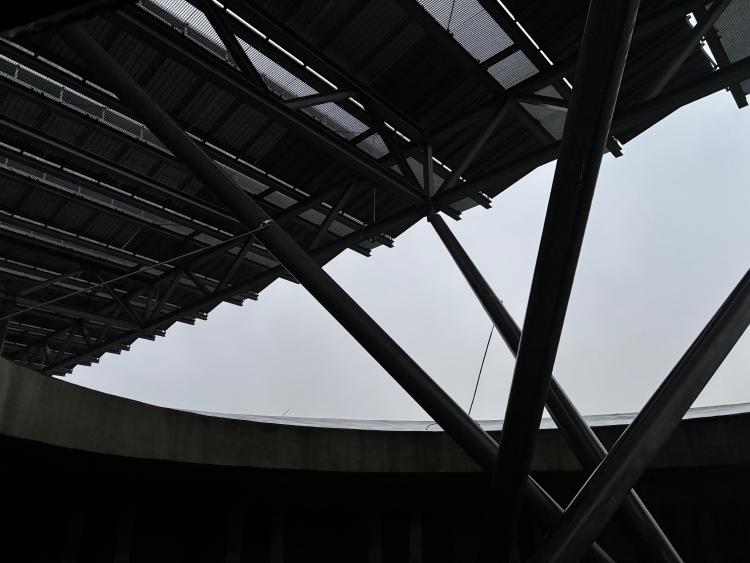 An almost black and white photo of 4 large angled poles emerging from inside a circular concrete structure supporting a louvred roof of solar panels.