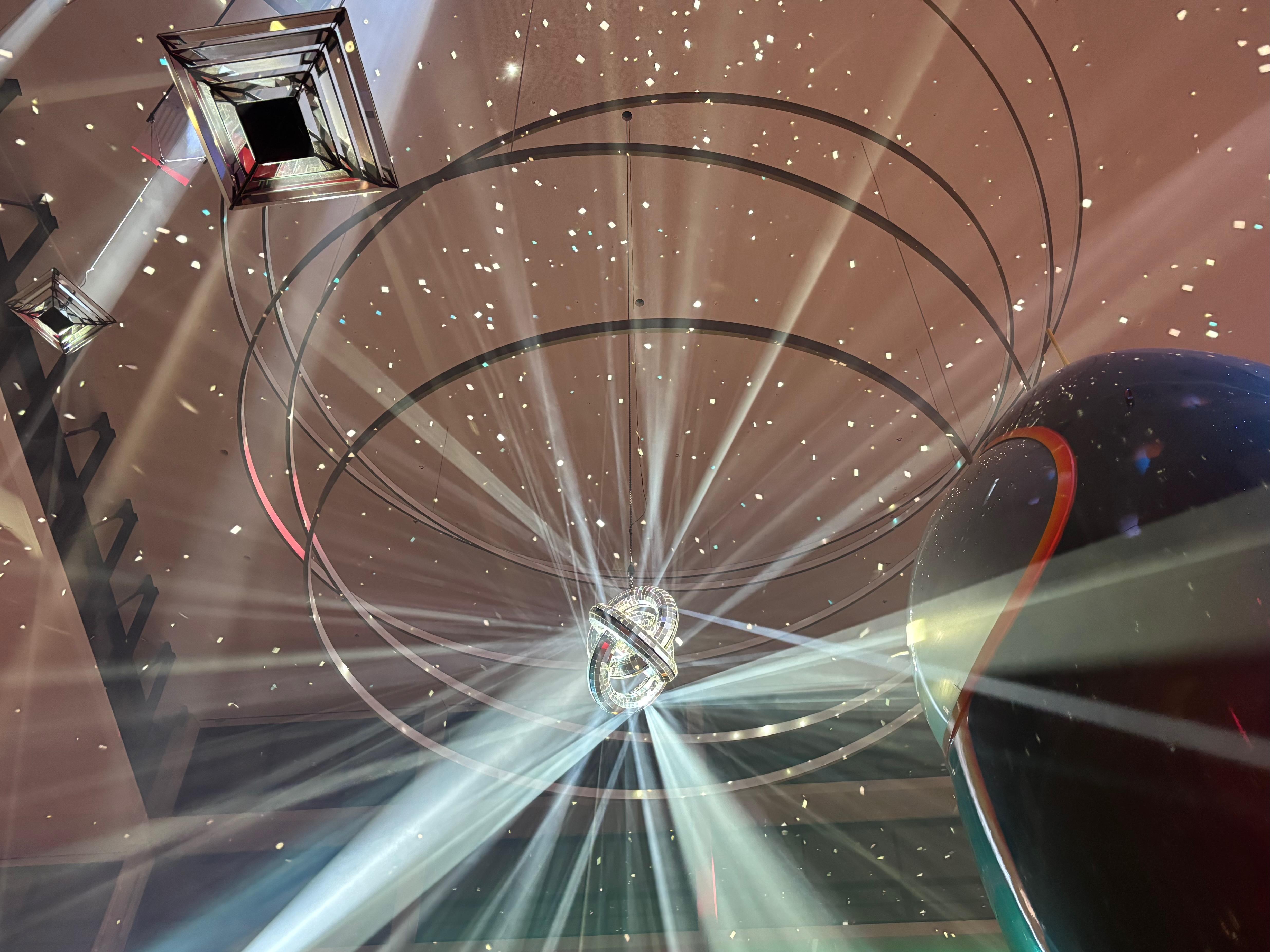 A "disco ball" made of three interlocking but not touching rings covered in mirrors. It hangs in a large lobby. A ton of beams of light are reflecting off of it. A stylised rocket is visible on the left of the frame.